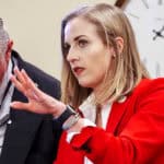 Allie Friesen, in a red suit and tie, is talking to a man in the Oklahoma Legislature.