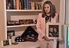 Woman in a pink suit holds a framed photo of a couple while standing beside a shelf of books and military memorabilia.