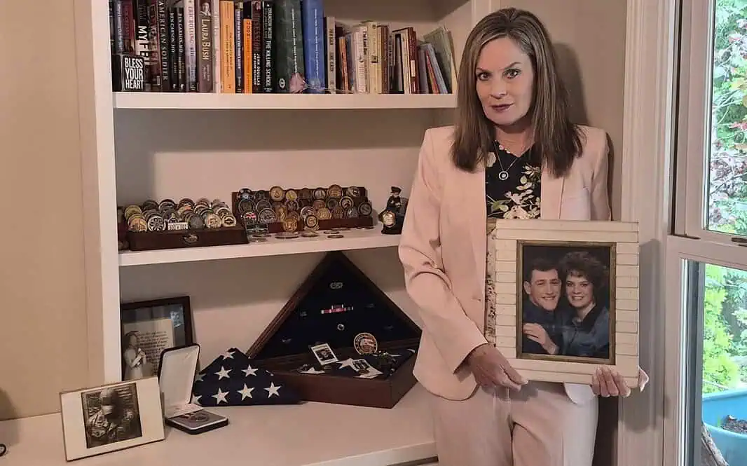 6165-1536x1235 Woman in a pink suit holds a framed photo of a couple while standing beside a shelf of books and military memorabilia.