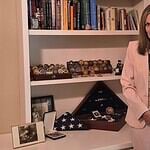 Woman in a pink suit holds a framed photo of a couple while standing beside a shelf of books and military memorabilia.