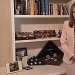 Gold Star Families Monument to be placed at Oklahoma Capitol Woman in a pink suit holds a framed photo of a couple while standing beside a shelf of books and military memorabilia.