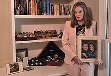 Woman in a pink suit holds a framed photo of a couple while standing beside a shelf of books and military memorabilia.