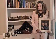 Gold Star Families Monument to be placed at Oklahoma Capitol Woman in a pink suit holds a framed photo of a couple while standing beside a shelf of books and military memorabilia.
