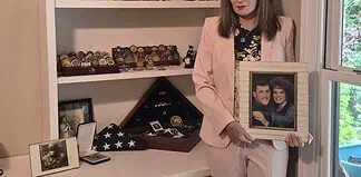 Woman in a pink suit holds a framed photo of a couple while standing beside a shelf of books and military memorabilia.