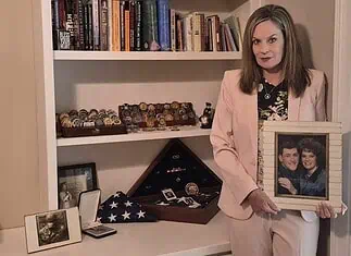 Gold Star Families Monument to be placed at Oklahoma Capitol Woman in a pink suit holds a framed photo of a couple while standing beside a shelf of books and military memorabilia.