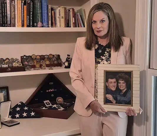 Gold Star Families Monument to be placed at Oklahoma Capitol Woman in a pink suit holds a framed photo of a couple while standing beside a shelf of books and military memorabilia.