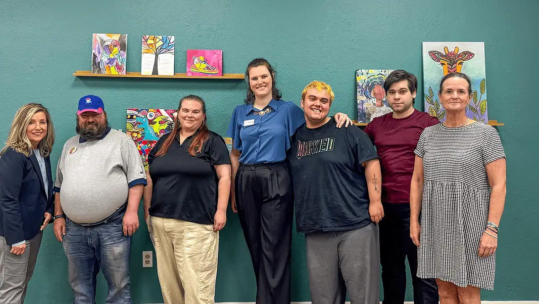 AutismOK Delta Dental Eight people pose for a group photo in an art studio with colorful paintings on a teal wall behind them.