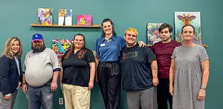 Eight people pose for a group photo in an art studio with colorful paintings on a teal wall behind them.