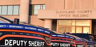 Row of black Dodge deputy sheriff patrol cars lined up in front of the Cleveland County Office Building exterior, displaying bold ‘DEPUTY SHERIFF’ lettering.