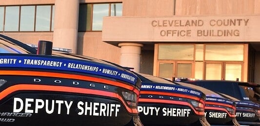 Row of black Dodge deputy sheriff patrol cars lined up in front of the Cleveland County Office Building exterior, displaying bold ‘DEPUTY SHERIFF’ lettering.