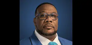 Portrait of a Black man in a blue suit, white shirt, and turquoise patterned tie, against a dark blue gradient background.