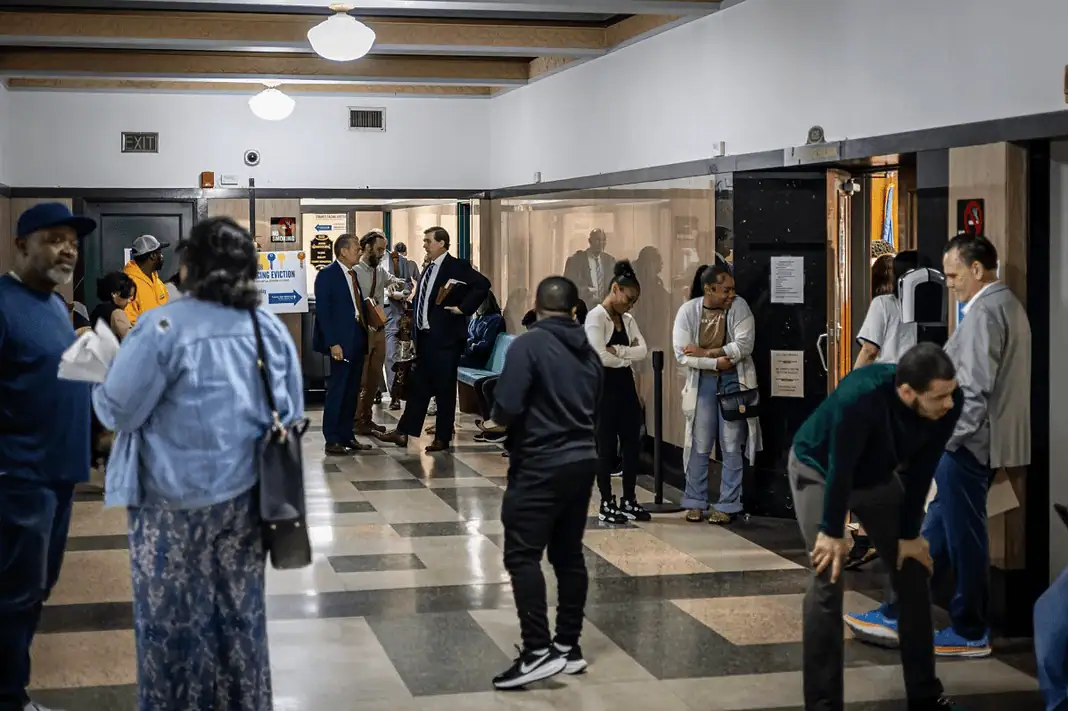 Eviction Court - Oklahoma Watch Diverse adults waiting in a hospital corridor, some talking near doors and seating area under bright lights in a medical facility.