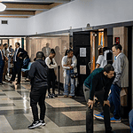 Diverse adults waiting in a hospital corridor, some talking near doors and seating area under bright lights in a medical facility.