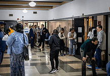 Diverse adults waiting in a hospital corridor, some talking near doors and seating area under bright lights in a medical facility.