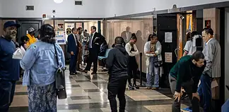 Diverse adults waiting in a hospital corridor, some talking near doors and seating area under bright lights in a medical facility.