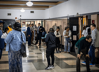 Diverse adults waiting in a hospital corridor, some talking near doors and seating area under bright lights in a medical facility.