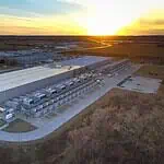 City Council puts hold on data centers to study electricity, water use Aerial view of a large industrial warehouse complex at sunset, with rows of equipment along the side and a river nearby.