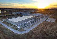 City Council puts hold on data centers to study electricity, water use Aerial view of a large industrial warehouse complex at sunset, with rows of equipment along the side and a river nearby.