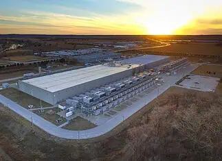 City Council puts hold on data centers to study electricity, water use Aerial view of a large industrial warehouse complex at sunset, with rows of equipment along the side and a river nearby.