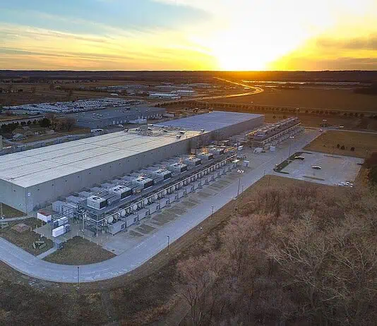 City Council puts hold on data centers to study electricity, water use Aerial view of a large industrial warehouse complex at sunset, with rows of equipment along the side and a river nearby.