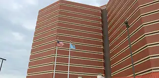 Tall red-brick building with beige horizontal bands, two flagpoles (American flag and blue flag) in front, and a parking lot with cars nearby.