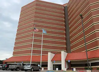 County cuts jail monitoring staff, meaning fewer eyes on detainees Tall red-brick building with beige horizontal bands, two flagpoles (American flag and blue flag) in front, and a parking lot with cars nearby.
