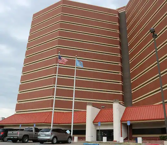 County cuts jail monitoring staff, meaning fewer eyes on detainees Tall red-brick building with beige horizontal bands, two flagpoles (American flag and blue flag) in front, and a parking lot with cars nearby.