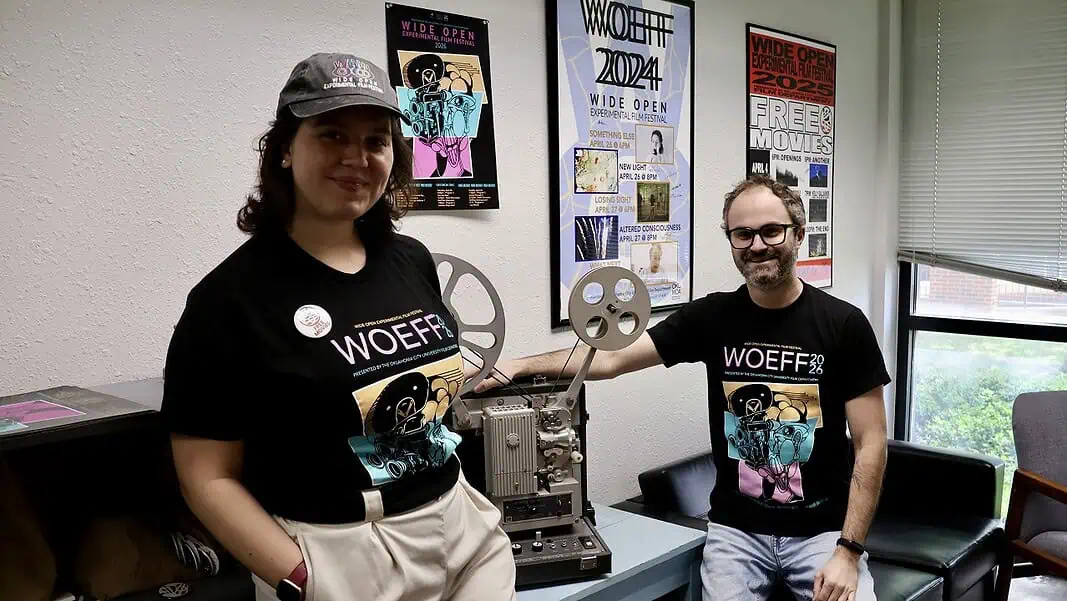 IMG_6894 Two people in WoEFF shirts stand beside a vintage film projector in a poster-filled room with festival posters on the wall to the right and left of them and a window nearby.