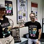Wide Open Experimental Film Fest spotlighting students, strangeness Two people in WoEFF shirts stand beside a vintage film projector in a poster-filled room with festival posters on the wall to the right and left of them and a window nearby.