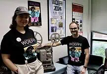 Wide Open Experimental Film Fest spotlighting students, strangeness Two people in WoEFF shirts stand beside a vintage film projector in a poster-filled room with festival posters on the wall to the right and left of them and a window nearby.