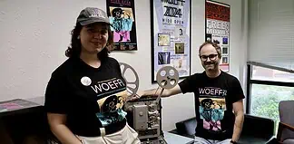 Two people in WoEFF shirts stand beside a vintage film projector in a poster-filled room with festival posters on the wall to the right and left of them and a window nearby.