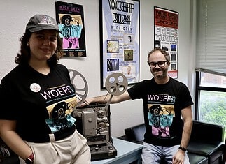 Two people in WoEFF shirts stand beside a vintage film projector in a poster-filled room with festival posters on the wall to the right and left of them and a window nearby.