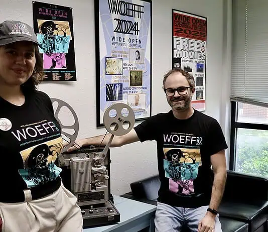 Wide Open Experimental Film Fest spotlighting students, strangeness Two people in WoEFF shirts stand beside a vintage film projector in a poster-filled room with festival posters on the wall to the right and left of them and a window nearby.