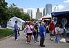 People walk through an outdoor street fair with white tents and vendors, city skyscrapers in the background.