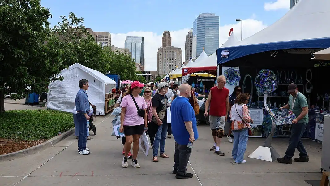 IMG_7753 People walk through an outdoor street fair with white tents and vendors, city skyscrapers in the background.