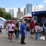 People walk through an outdoor street fair with white tents and vendors, city skyscrapers in the background.