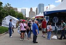 People walk through an outdoor street fair with white tents and vendors, city skyscrapers in the background.