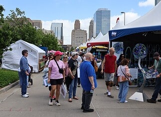 People walk through an outdoor street fair with white tents and vendors, city skyscrapers in the background.