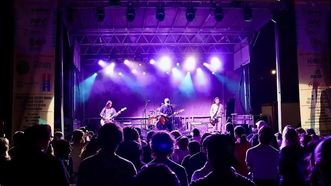 IMG_8430 Band performs on a lit stage with purple and blue lights as a crowd watches from the foreground at a nighttime concert.