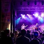 Band performs on a lit stage with purple and blue lights as a crowd watches from the foreground at a nighttime concert.