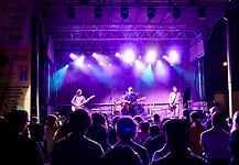 Band performs on a lit stage with purple and blue lights as a crowd watches from the foreground at a nighttime concert.