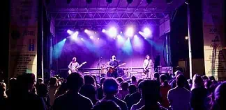 Band performs on a lit stage with purple and blue lights as a crowd watches from the foreground at a nighttime concert.