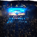Aerial view of a nighttime outdoor concert with a large crowd facing a brightly lit stage and blue stage lights.