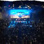 Here’s your guide to the can’t-miss acts at Norman Music Fest 2026 Aerial view of a nighttime outdoor concert with a large crowd facing a brightly lit stage and blue stage lights.