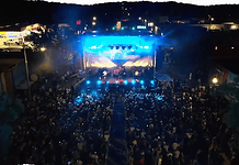 Aerial view of a nighttime outdoor concert with a large crowd facing a brightly lit stage and blue stage lights.
