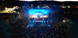 Aerial view of a nighttime outdoor concert with a large crowd facing a brightly lit stage and blue stage lights.