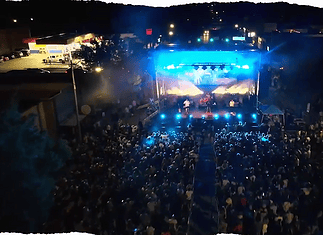 Aerial view of a nighttime outdoor concert with a large crowd facing a brightly lit stage and blue stage lights.