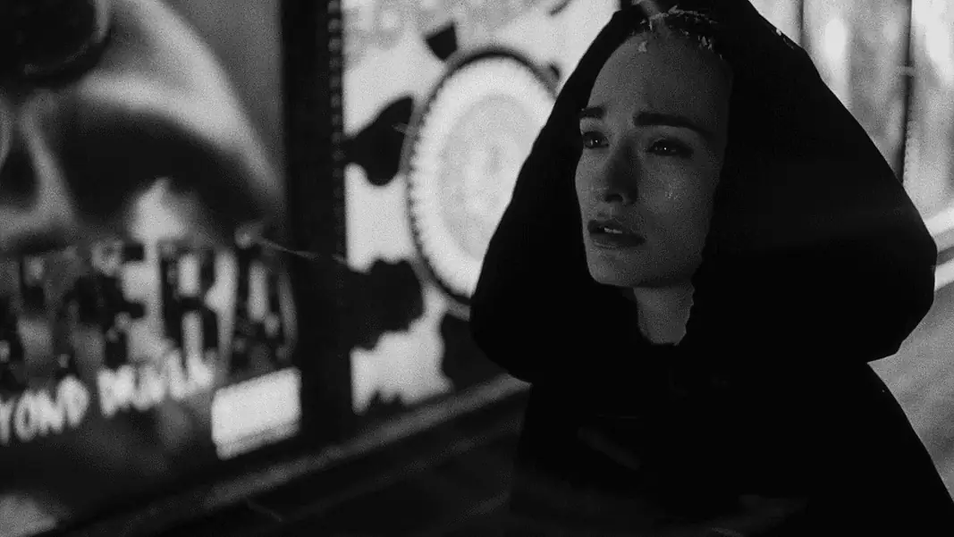 Nadja Woman in a dark hooded coat stands in a dim urban street at night, looking worried or sad.