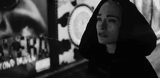 Woman in a dark hooded coat stands in a dim urban street at night, looking worried or sad.