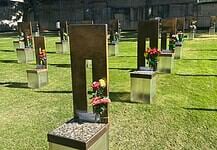 Rows of bronze and metal grave markers with colorful flowers on a well-kept grassy cemetery lawn.
